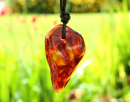 A teardrop-shaped amber glass pendant necklace with a black cord, shown hanging against a blurred green garden background.