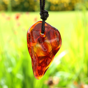 A teardrop-shaped amber glass pendant necklace with a black cord, shown hanging against a blurred green garden background.