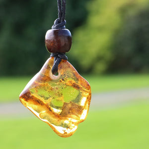 A raw, irregular amber glass pendant with dark wood bead on a black cord, photographed outdoors against a blurred green park background.