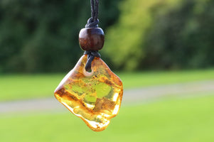 A raw, irregular amber glass pendant with dark wood bead on a black cord, photographed outdoors against a blurred green park background.