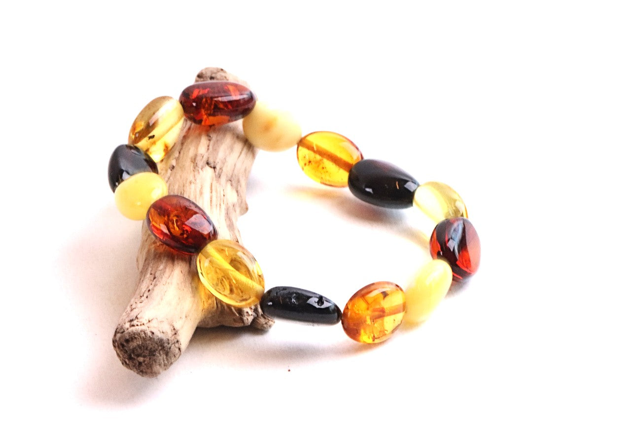 A beaded bracelet made of polished oval amber-like beads in yellow, orange, and black, displayed on a small piece of driftwood against a white background.