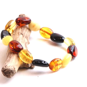 A beaded bracelet made of polished oval amber-like beads in yellow, orange, and black, displayed on a small piece of driftwood against a white background.