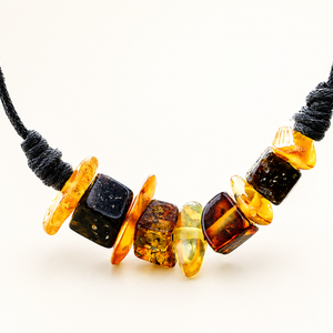 A close-up of a necklace with a black cord and a row of polished amber beads in various shapes and colors, including yellow, orange, and dark brown, on a white background.