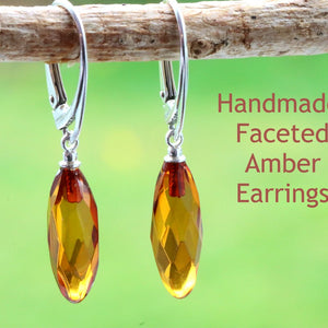 A pair of handmade faceted amber earrings hanging from a wooden branch, with a blurred green background.