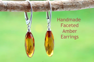 A pair of handmade faceted amber earrings hanging from a wooden branch, with a blurred green background.