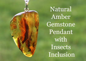 A teardrop-shaped natural amber gemstone pendant with insect inclusion, suspended by a silver loop, against an outdoor background.