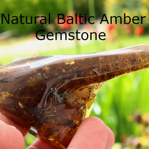 A hand holding a polished natural Baltic amber gemstone against a blurred green garden background, with the text 'Natural Baltic Amber Gemstone' overlaid at the top.
