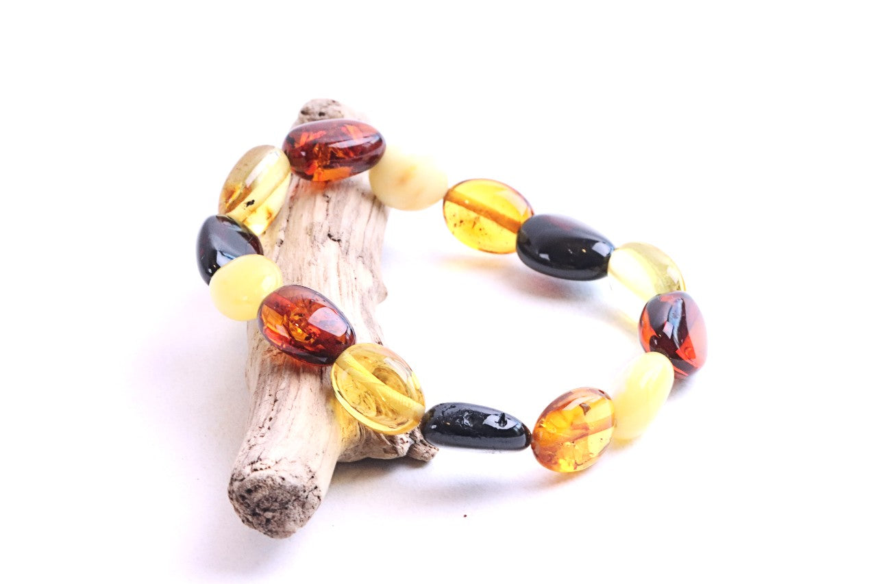 A beaded bracelet made of polished amber and glass beads in yellow, orange, and black, displayed on a small piece of driftwood against a white background.