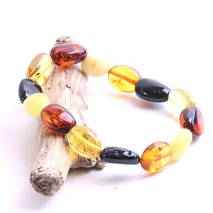 A beaded bracelet made of polished amber and glass beads in yellow, orange, and black, displayed on a small piece of driftwood against a white background.