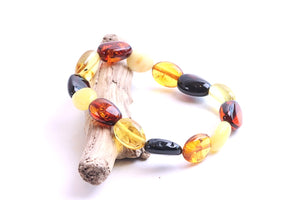 A beaded bracelet made of polished amber and glass beads in yellow, orange, and black, displayed on a small piece of driftwood against a white background.