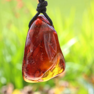 A polished amber-like stone pendant with a black cord, photographed outdoors against a blurred green grassy background.