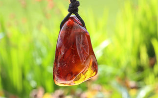 A polished amber-like stone pendant with a black cord, photographed outdoors against a blurred green grassy background.