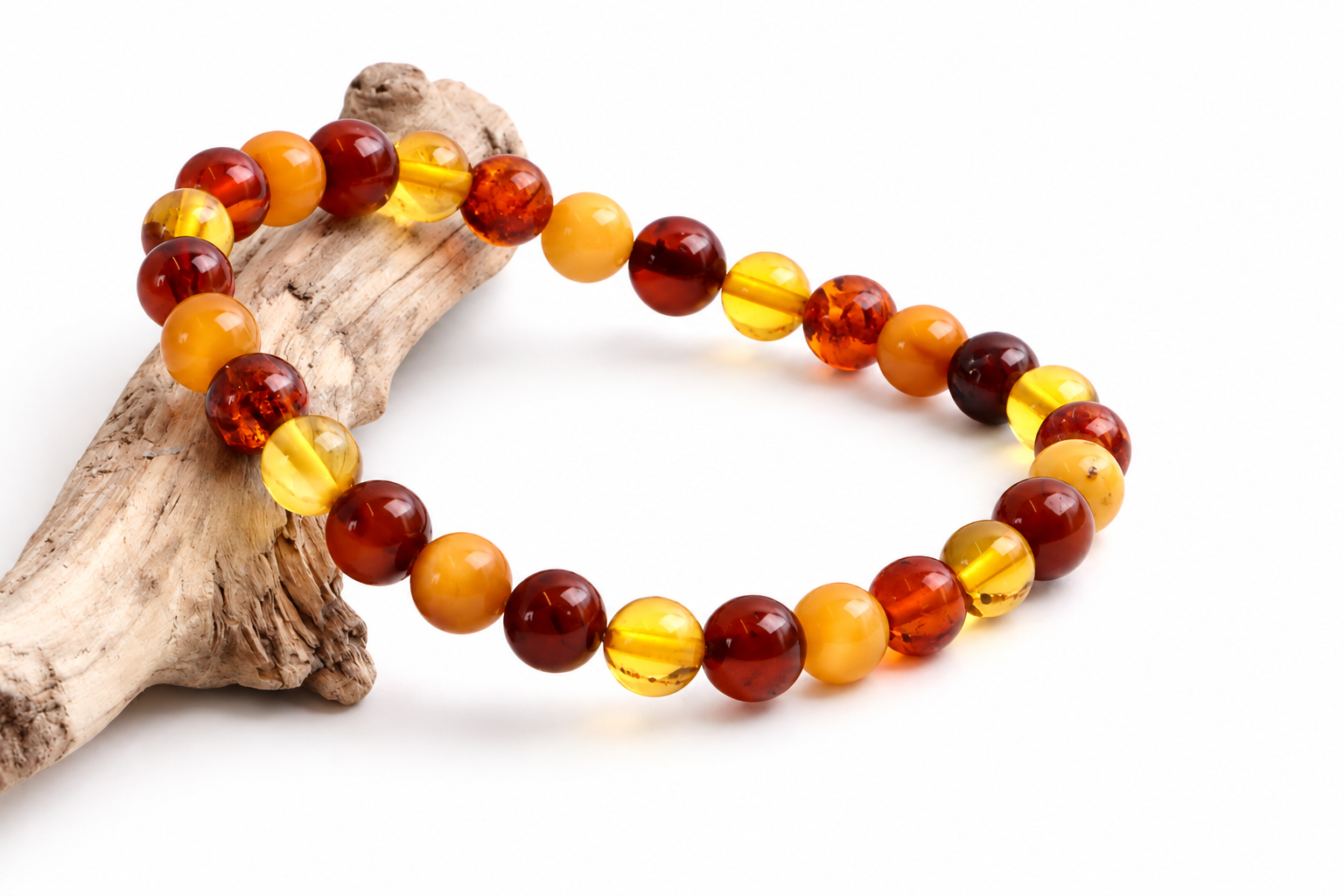 A beaded bracelet made of polished round amber-like beads in translucent yellow, orange, and deep reddish brown, arranged on a piece of driftwood against a white background.