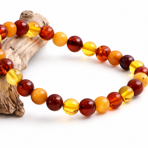 A beaded bracelet made of polished round amber-like beads in translucent yellow, orange, and deep reddish brown, arranged on a piece of driftwood against a white background.
