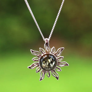 A silver sun pendant necklace with a central round smoky gemstone, hanging against a blurred green outdoor background.