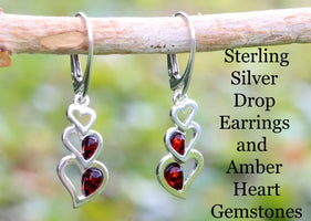 A pair of sterling silver drop earrings with red amber heart-shaped gemstones, displayed on a wooden branch against a blurred green background.