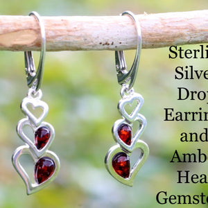 A pair of sterling silver drop earrings with red amber heart-shaped gemstones, displayed on a wooden branch against a blurred green background.