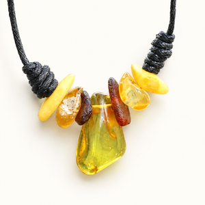 A handmade tumbled stone necklace featuring a large central yellow-green amber bead, flanked by smaller yellow, orange, and brown stones, suspended on a black cord against a white background.